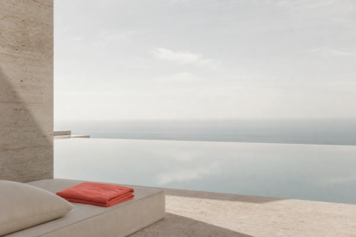 The edge of a minimalist villa where an infinity pool meets a hazy sky, a lounger with a single folded coral towel